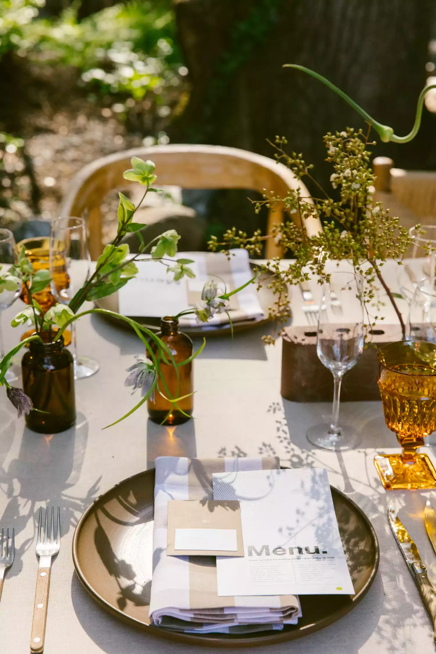 Close Up of Reception Table Setting With Simple Flowers and Leaves in Brown Bottles, Gold tinted Glasses, and Menu On Top of Brown and White Checkered Napkin and Brown Plate