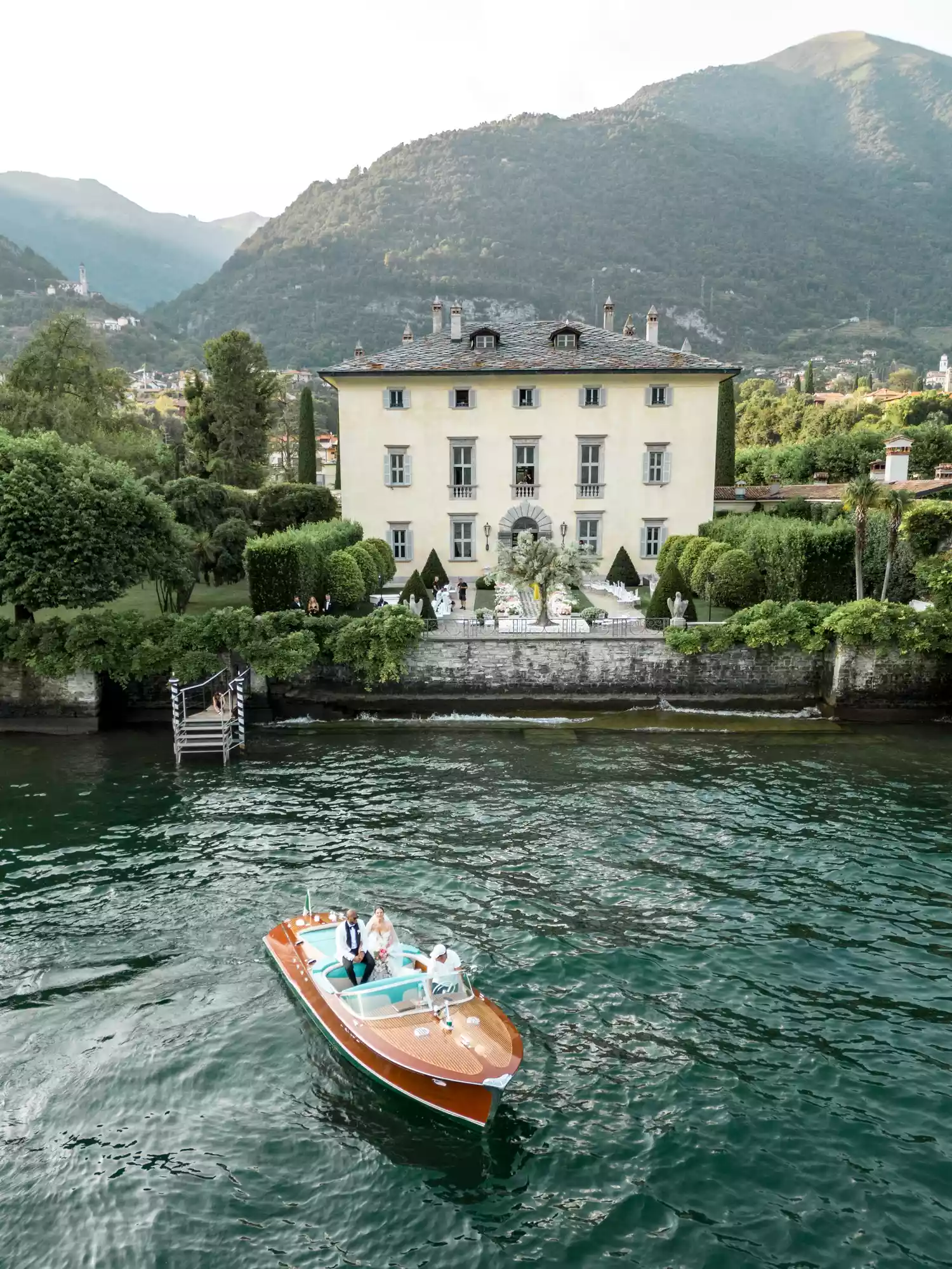 Bride and groom riding on a wooden boat on lake Como with their villa venue in the background