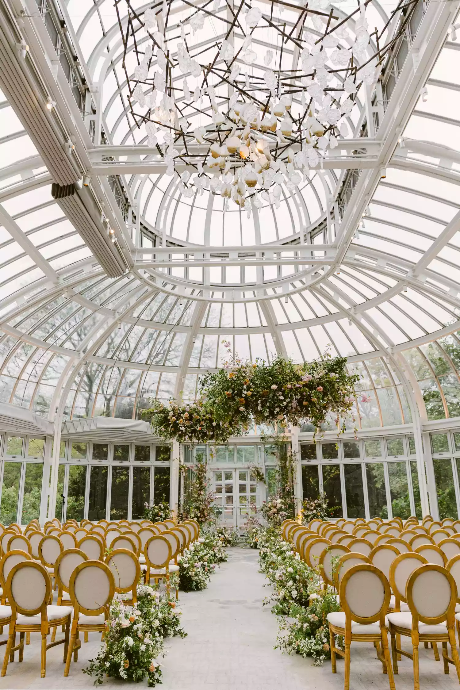 greenhouse wedding venue with gold and white ceremony chairs and a large hanging flower installation