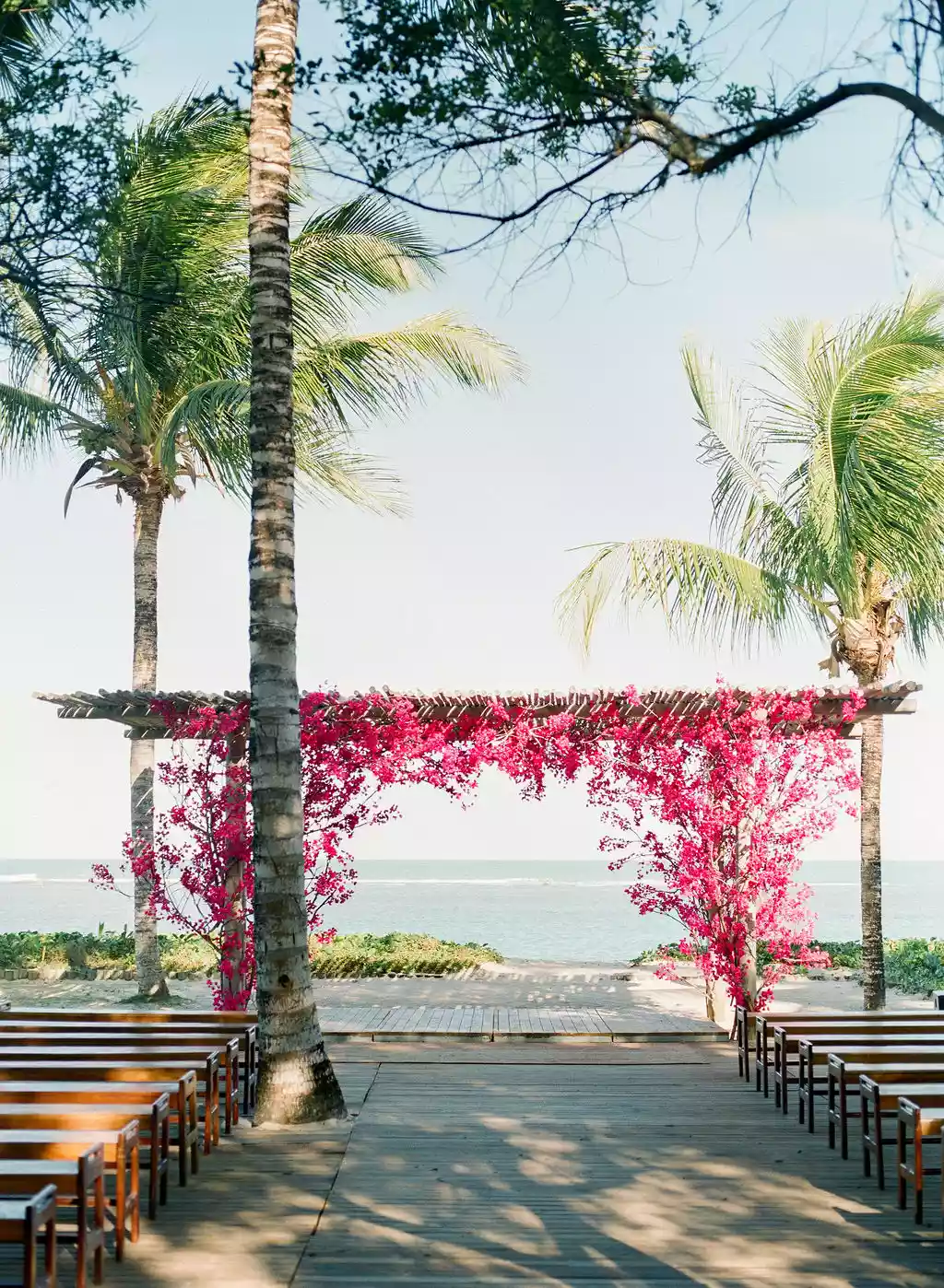 Wedding ceremony setup at Brazilian beach with benches, a wooden altar decorated with bougainvillea, and palm trees