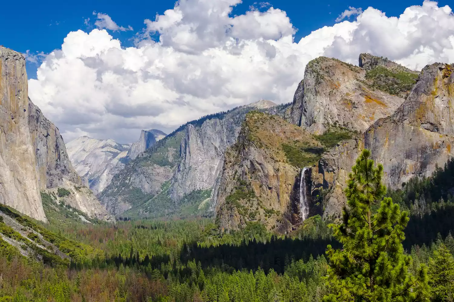 A view of mountains and vibrant green trees in front of clouds and blue sky at Bridal Veil Falls in Yosemite National Park in California.