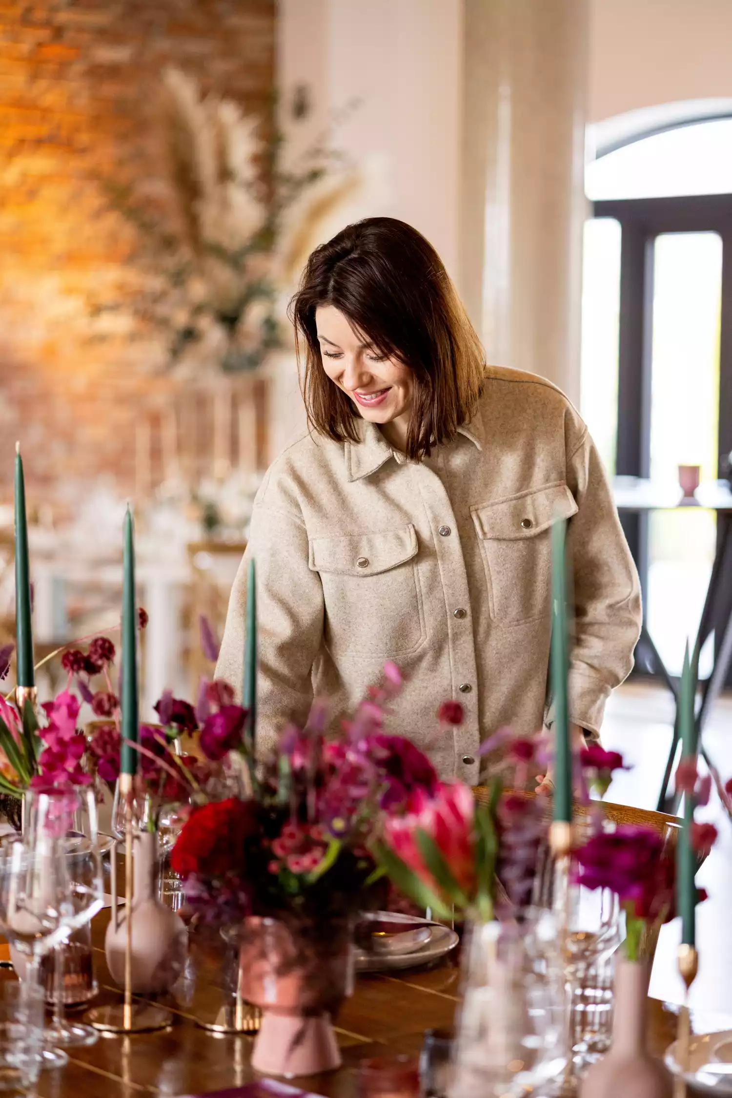 Woman With Dark, Short Hair Smiling and Standing Over Pink Floral Centerpiece and Green Taper Candles