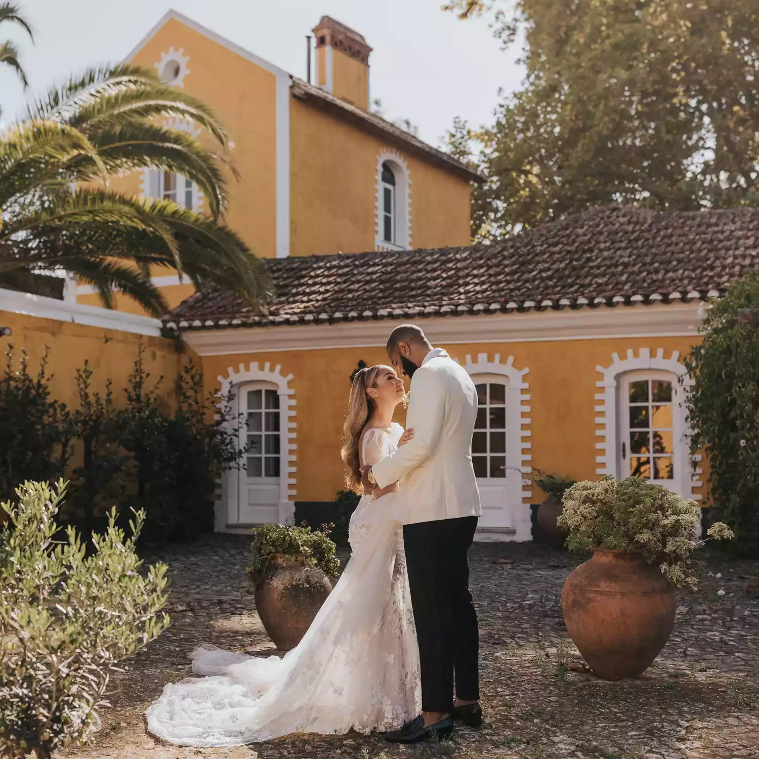 A bride and groom wearing white wedding attire, embracing in front of an orange-painted wedding venue with lush vegetation during a daytime wedding.