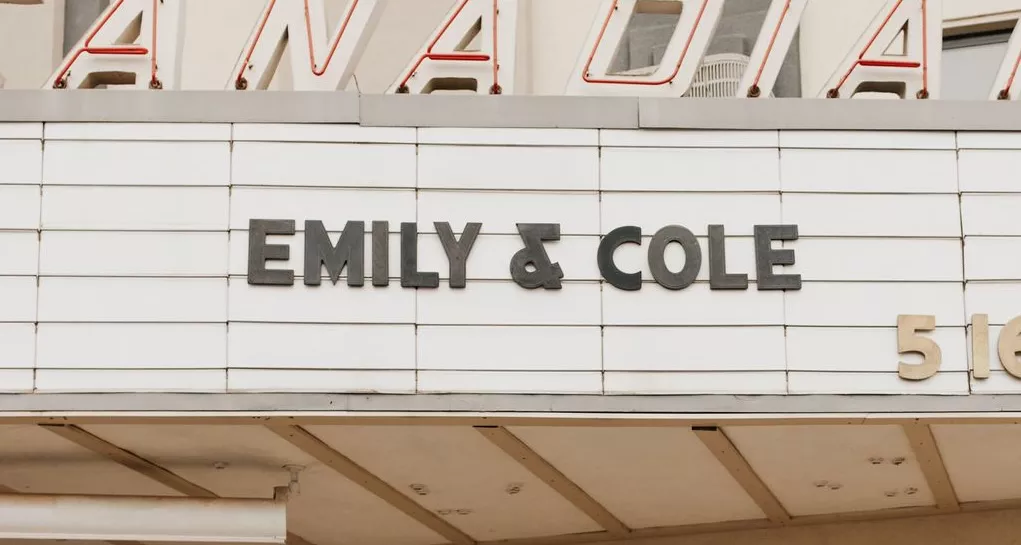 Bride and groom kissing under theater marquee sign that says "Emily & Cole" during a theater wedding.