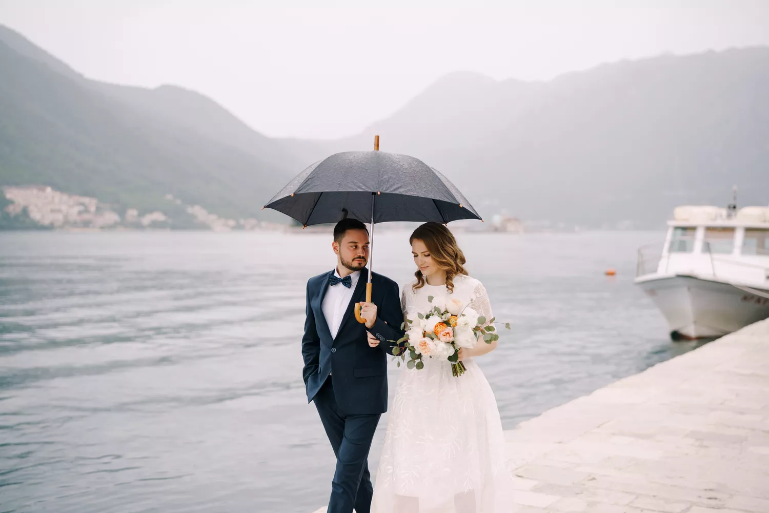 Bride and groom holding an umbrella while walking along the water on rainy wedding day