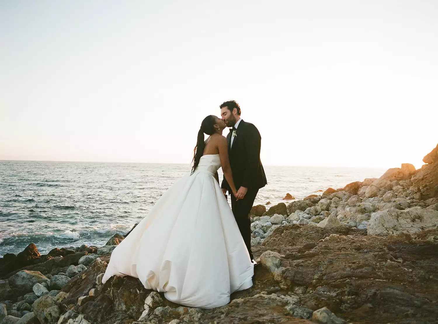 bride and groom kissing on rocks near the ocean