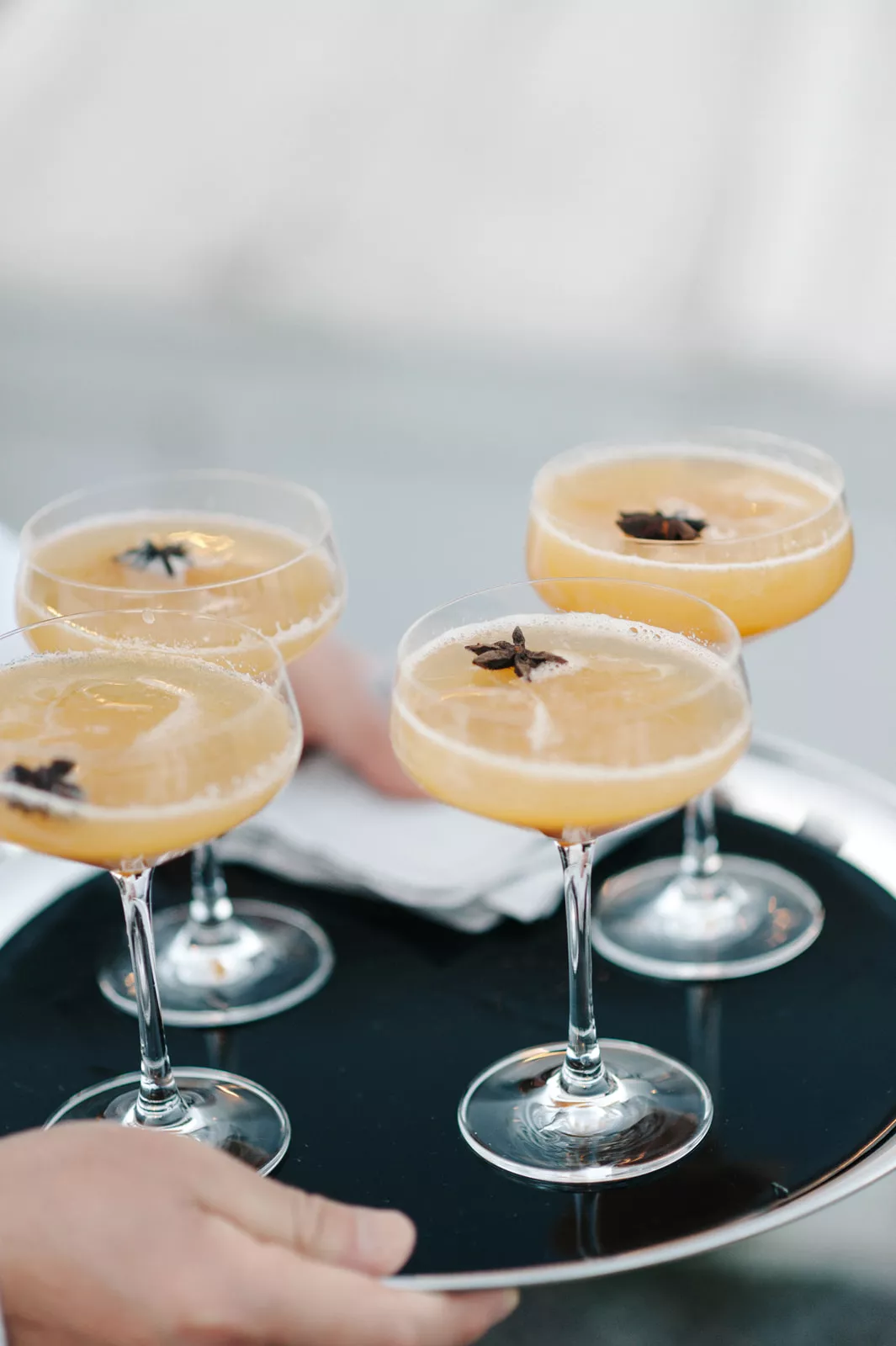 Four orange cocktails with garnish in couple glasses being served by a caterer at an outdoor wedding.